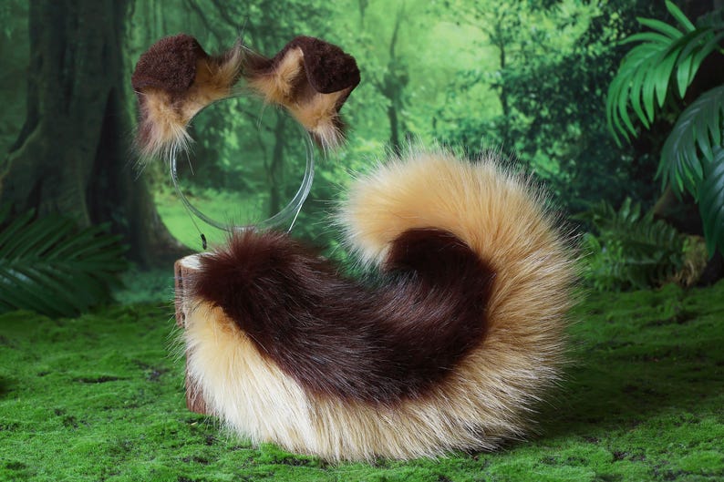 Pode incluir: Um conjunto de orelhas de animais de pele sint&eacute;tica castanha e bege numa bandolete e uma cauda a condizer. A cauda &eacute; uma mistura de pele castanha, bege e creme. Os artigos s&atilde;o exibidos numa superf&iacute;cie verde com um fundo de floresta.