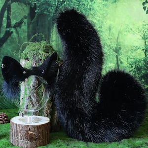 Puede incluir: Un conjunto de cola y diadema de orejas de piel sintética negra. La cola es grande y esponjosa, y las orejas están unidas a una diadema transparente. El conjunto se exhibe sobre una base de madera con un fondo de bosque.