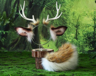May include: A deer-themed costume set featuring a headband with white antlers and faux fur ears, and a matching fluffy tail. The set is displayed on a wooden stand against a backdrop of lush greenery.