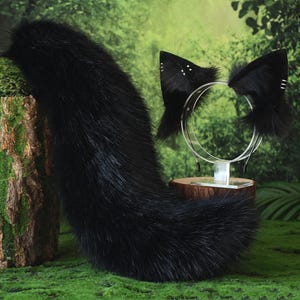 May include: A black faux fur tail and cat ear headband set. The tail is long and fluffy. The headband features black cat ears with white accents. The items are displayed on a green mossy surface.