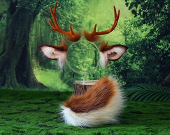 May include: A woodland-themed costume set featuring brown faux fur deer ears, antlers, and a tail. The ears and tail have white accents. The antlers are a reddish-brown color. The set is displayed on a green mossy surface.