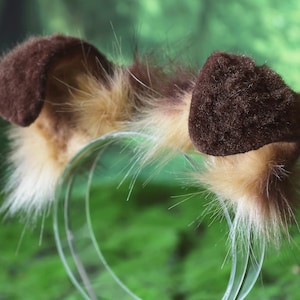 Pode incluir: Um par de acess&oacute;rios de faixa de orelha de cachorro marrons. As orelhas s&atilde;o de cor marrom escuro e t&ecirc;m uma textura macia e peluda. A faixa de cabe&ccedil;a &eacute; de pl&aacute;stico transparente. As orelhas s&atilde;o cercadas por pele marrom e branca.