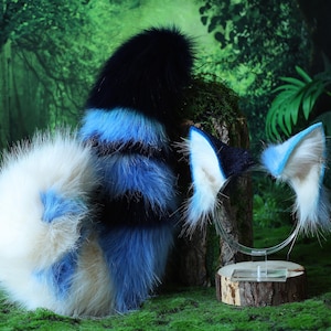 May include: A furry cosplay set featuring a tail and a headband with ears. The tail is white, blue, and black. The ears are white and light blue. The items are displayed on a green surface with a forest backdrop.