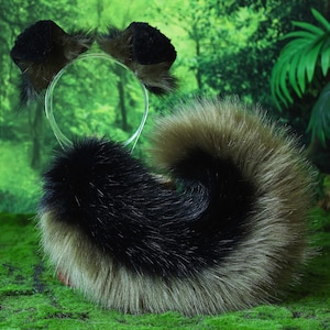 May include: A costume set with a headband and tail. The headband has two small, dark brown ears. The tail is black, brown, and tan, and is curled. The set is designed to resemble an animal. The background is a green, forest-like scene.