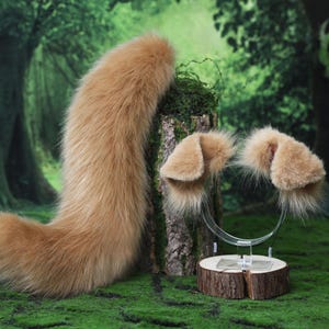 May include: A long, fluffy, light brown faux fur tail positioned next to a pair of matching light brown faux fur animal ears attached to a clear headband. The tail and ears feature a soft, dense texture with a natural, tapered shape.