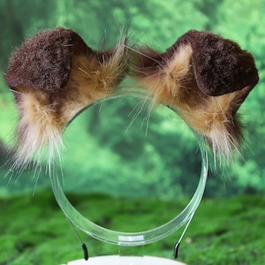Pode incluir: Uma tiara com duas orelhas de cachorro de pel&uacute;cia. As orelhas s&atilde;o marrons com detalhes em pele bronzeada e branca. A tiara &eacute; de pl&aacute;stico transparente. O fundo &eacute; verde desfocado.