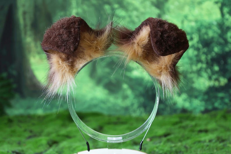 Pode incluir: Uma tiara com duas orelhas de cachorro de pel&uacute;cia. As orelhas s&atilde;o marrons com detalhes em pele bronzeada e branca. A tiara &eacute; de pl&aacute;stico transparente. O fundo &eacute; verde desfocado.