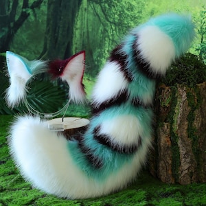 May include: A fluffy, faux fur tail and ear set in white, teal, and dark brown. The tail is striped and the ears are attached to a clear headband. The set is displayed on a mossy surface with a forest background.