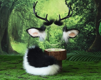 May include: A deer costume set featuring a black and white faux fur tail, a headband with black antlers, and matching ear accents. The ears have a pinkish-tan interior and white fur trim. The set is displayed on a wooden stand against a forest backdrop.