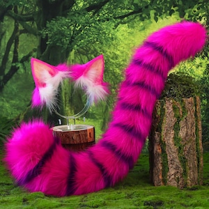 Op de afbeelding: Een set bestaande uit een paar kattenoren van imitatiebont op een haarband en een lange, bijpassende staart van imitatiebont. Beide items hebben een felroze kleur met donkerpaarse horizontale strepen en witte accenten in de oren.
