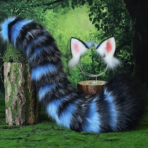 May include: A faux fur cosplay set featuring a striped blue and black tail and a headband with white and pink cat ears. The tail is thick and fluffy, and the ears have pink inner details. The set is displayed on a mossy surface.