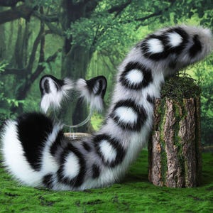 May include: A set of faux fur animal ears and a long, curved tail featuring a white base with black and grey circular spots. The ears are upright with black inner details, and the long, plush tail has a distinct pattern of dark spots.