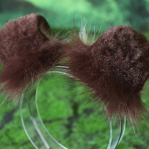 Pode incluir: Um par de orelhas de animais castanhas e fofas em uma tiara transparente. As orelhas s&atilde;o de cor castanha escura com uma apar&ecirc;ncia macia e texturizada e pelos longos e finos. A tiara &eacute; feita de pl&aacute;stico transparente.