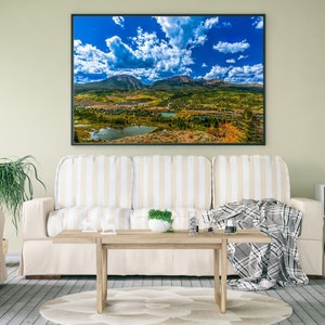 Pode incluir: Uma fotografia de paisagem emoldurada de uma cordilheira com um céu azul e nuvens brancas. A fotografia está pendurada em uma parede acima de um sofá listrado branco com uma manta e uma mesa de centro de madeira.
