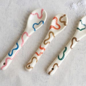May include: Three off-white ceramic spoons featuring hand-painted wavy lines in various colors. The first spoon has pink and blue lines, the second has orange and brown lines, and the third has green and brown lines. Each handle has a small hole.