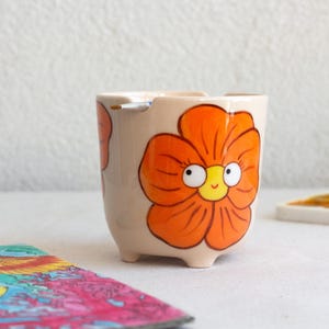 以下が含まれることがあります： オレンジ色の花の模様が描かれた、小さなベージュ色の陶器の植木鉢。花は白い目と黄色の中心を持つ笑顔の顔をしています。植木鉢には3つの小さな足があり、白い表面に置かれています。