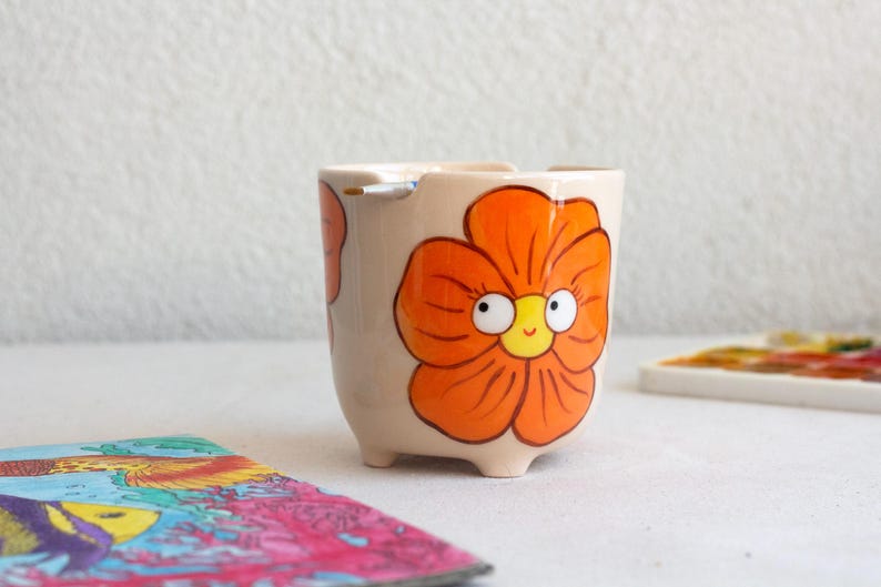 以下が含まれることがあります： オレンジ色の花の模様が描かれた、小さなベージュ色の陶器の植木鉢。花は白い目と黄色の中心を持つ笑顔の顔をしています。植木鉢には3つの小さな足があり、白い表面に置かれています。