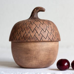 Puede incluir: Un recipiente de cerámica marrón con forma de bellota, con una tapa texturizada que presenta un patrón geométrico y una base lisa. La tapa tiene un tallo curvado en la parte superior. Tres cerezas de color rojo oscuro se sitúan a la derecha.
