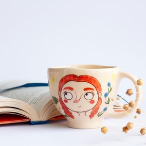 May include: A white ceramic mug with a hand-painted design of a woman with red hair and blue eyes. The mug has a handle and is decorated with yellow and blue flowers.