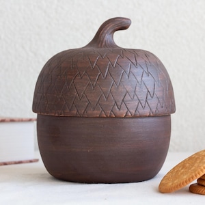 May include: A dark brown ceramic jar shaped like an acorn with a removable lid. The lid features a carved, repeating triangular pattern and a curved stem at the top. The base of the jar is smooth and rounded, providing a simple, functional container.