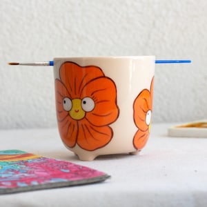 以下が含まれることがあります： オレンジ色の花のイラストと青い筆が描かれた、クリーム色の陶器の鉢。 鉢には小さな足が3つあり、漫画風の花で飾られています。 カラフルなコースターと絵の具のパレットが背景にあります。