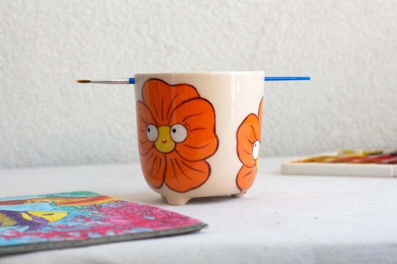 以下が含まれることがあります： オレンジ色の花のイラストと青い筆が描かれた、クリーム色の陶器の鉢。 鉢には小さな足が3つあり、漫画風の花で飾られています。 カラフルなコースターと絵の具のパレットが背景にあります。
