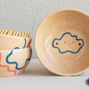 Puede incluir: Un conjunto de cuencos de cerámica beige con diseños abstractos azules y rosas. Un cuenco presenta un diseño de nube sonriente con contorno azul y puntos rojos. Los cuencos están moteados y tienen un aspecto rústico.
