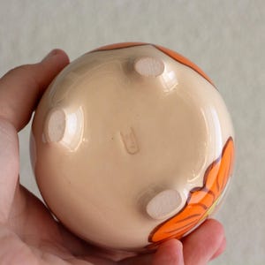 以下が含まれることがあります： ベージュのベースとオレンジ色の花の模様が施された、小さくて丸いセラミックの花瓶。花瓶には3つの小さな足があり、手で持たれています。花瓶の底には小さなスタンプがあります。