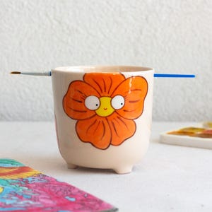 以下が含まれることがあります： クリーム色のセラミックカップ。オレンジ色の花の模様が描かれています。カップには3つの小さな足があり、両側に筆が置かれています。カラフルな絵と水彩パレットが背景にあります。