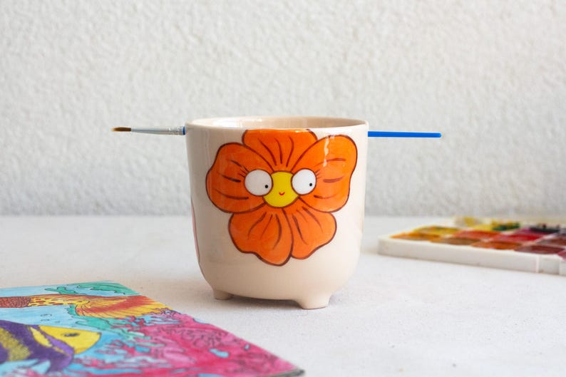 以下が含まれることがあります： クリーム色のセラミックカップ。オレンジ色の花の模様が描かれています。カップには3つの小さな足があり、両側に筆が置かれています。カラフルな絵と水彩パレットが背景にあります。