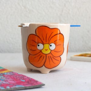 以下が含まれることがあります： クリーム色のセラミックカップに、大きなオレンジ色の花の模様。花は黄色の中心と漫画のような目があります。筆と青い棒が縁に置かれています。カラフルなイラストが前景にあります。