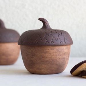Op de afbeelding: Twee eikelvormige keramische potten met donkerbruine, getextureerde deksels en lichtbruine onderkanten. De voorste pot heeft een geometrisch chevronpatroon in het deksel gegraveerd. Rechts liggen twee ronde koekjes met een laagje chocolade.