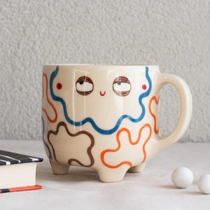 Puede incluir: Una taza de cerámica color crema con un rostro pintado a mano que muestra ojos mirando hacia arriba y una pequeña sonrisa roja. El exterior está decorado con líneas abstractas, onduladas y entrelazadas en azul, naranja y marrón, sobre tres patas.