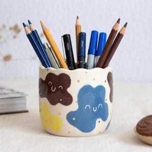 May include: A cylindrical ceramic pencil holder with a scalloped top edge, featuring a cream-colored base decorated with brown, blue, and yellow blob-shaped patterns, each with a simple smiling face, filled with various colored pencils and pens.