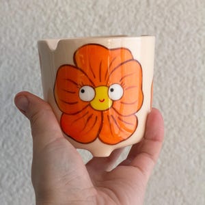 以下が含まれることがあります： 手描きのセラミック灰皿。オレンジ色の気まぐれな花の模様。花は黄色の中心と漫画の目があります。灰皿はベージュで、タバコ用の小さな切り込みがあります。