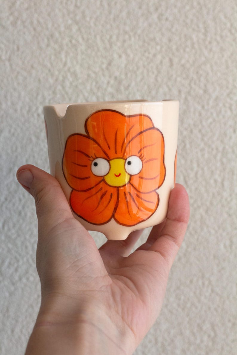 以下が含まれることがあります： 手描きのセラミック灰皿。オレンジ色の気まぐれな花の模様。花は黄色の中心と漫画の目があります。灰皿はベージュで、タバコ用の小さな切り込みがあります。