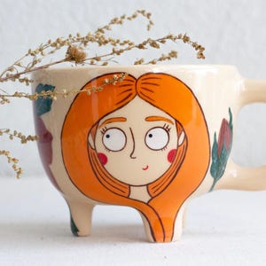 May include: Cream-colored ceramic mug with a whimsical design. The mug features a cartoon illustration of a woman with orange hair and red cheeks. The mug has three small feet and a rounded handle.