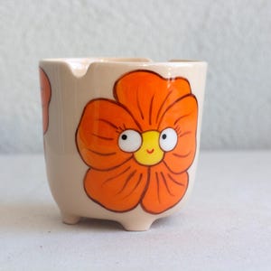 以下が含まれることがあります： ベージュ色の小さな陶器の灰皿。オレンジ色の花柄が描かれており、花には白い目と黄色の中心部を持つ笑顔の顔があります。灰皿には3つの小さな足が付いています。