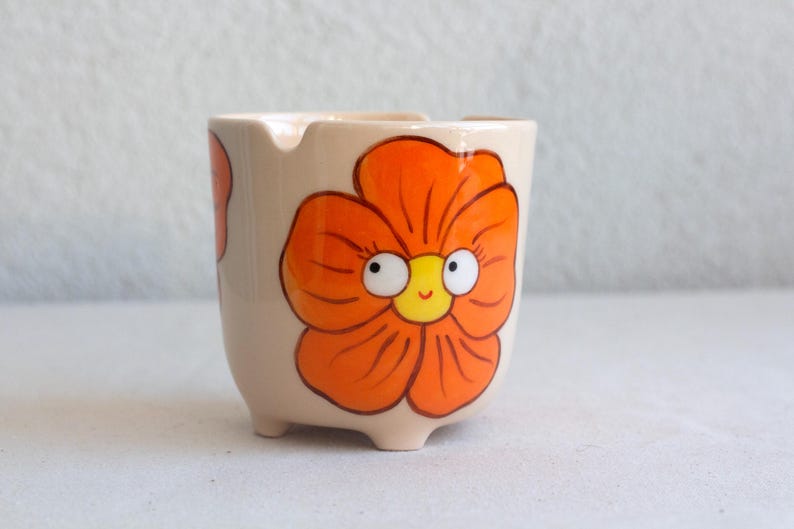 以下が含まれることがあります： ベージュ色の小さな陶器の灰皿。オレンジ色の花柄が描かれており、花には白い目と黄色の中心部を持つ笑顔の顔があります。灰皿には3つの小さな足が付いています。