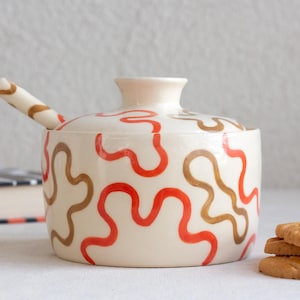 May include: A cream-coloured ceramic sugar bowl with a matching lid and a small spoon. The exterior of the bowl and the handle of the spoon feature a hand-painted pattern of wavy, irregular lines in vibrant orange and muted brown colours.