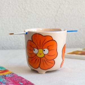 以下が含まれることがあります： 小さなオフホワイトのセラミックポット。オレンジ色の花のデザインが描かれており、花には大きな目と黄色の中心を持つ笑顔の顔があります。青い柄の筆が上に置かれています。小さなアイテムや植物を入れるためのポットです。
