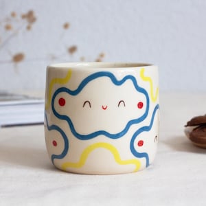 Puede incluir: Una taza de cerámica de color crema con un diseño de cara sonriente. La taza presenta líneas onduladas azules y amarillas y puntos rojos. La taza es cilíndrica. Galletas de chocolate en un plato pequeño a la derecha.