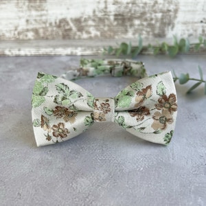 Op de afbeelding: Een bloemenstrik en bijpassende halsband. De strik heeft een crèmekleurige achtergrond met groene en bruine bloempatronen. De halsband en strik zijn gemaakt van stof.