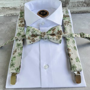 Op de afbeelding: Een wit overhemd met een bloemenstrik en bretels. De strik en bretels hebben een crèmekleurige achtergrond met groene en bruine bloempatronen. De bretels hebben metalen clips.