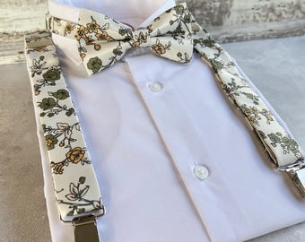 Puede incluir: Una camisa blanca con un conjunto de pajarita y tirantes florales. La pajarita y los tirantes presentan un fondo crema con un estampado de flores y hojas verdes y amarillas. Los tirantes tienen clips metálicos plateados.