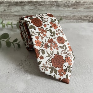 Può includere: Una cravatta bianca con un motivo floreale ripetuto in tonalità ruggine, arancione e verde. La cravatta è arrotolata, mostrando il design dettagliato dei fiori e delle foglie. Lo sfondo è una superficie grigia neutra.