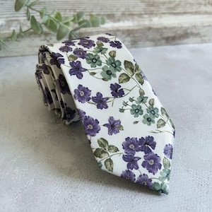 Peut inclure: Une cravate à motifs floraux, avec un fond blanc orné de fleurs violettes et vertes, ainsi que de feuilles vertes. La cravate est pliée, suggérant un tissu lisse. Le motif floral est uniformément réparti sur la cravate.