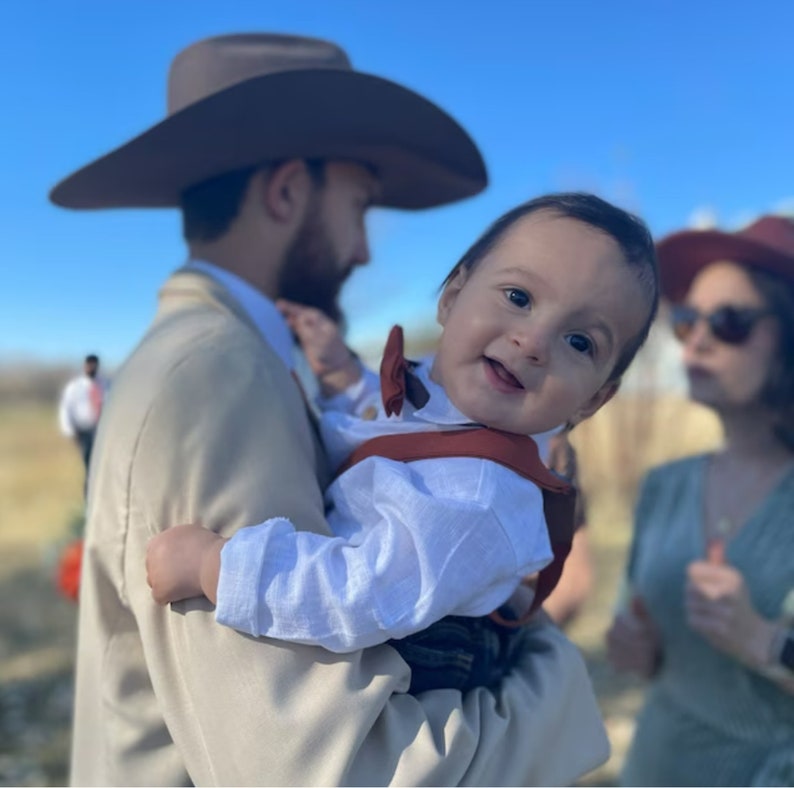 Op de afbeelding: Een man in een bruine cowboyhoed houdt een baby vast die een wit overhemd met een bruine strik en bretels draagt. De baby glimlacht en kijkt naar de camera. Een vrouw in een rood-witte gebloemde jurk staat achter hen.