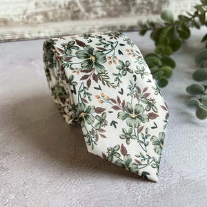 Könnte beinhalten: Eine gerollte Krawatte mit floralem Muster. Die Krawatte hat einen weißen Hintergrund, verziert mit einem Muster aus grünen Blumen, braunen Blättern und kleinen orangefarbenen Details. Der Stoff wirkt weich und eignet sich für formelle oder halbformelle Anlässe. Die Krawatte ist ca. 147 cm lang.