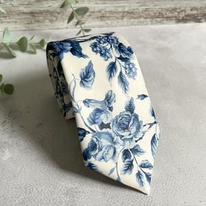 Könnte beinhalten: Eine gerollte Krawatte mit cremefarbenem Hintergrund und blauem Blumenmuster. Das Muster zeigt Rosen und andere Blumen mit Blättern. Die Krawatte ist aus glattem Stoff gefertigt und für formelle Anlässe geeignet.